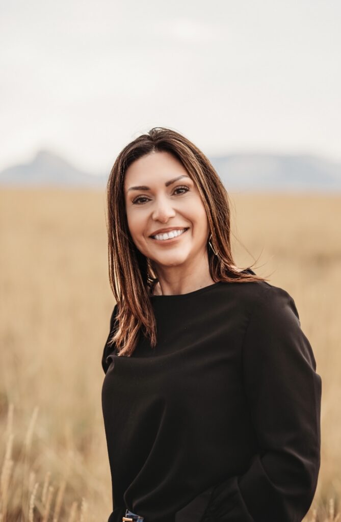 Close-up portrait of woman with a sincere smile, standing against a backdrop of tall grass and distant mountains.