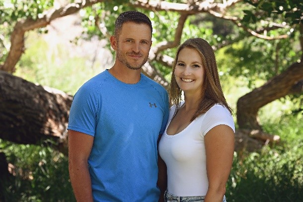 A close-up of Kimberly Spitzer standing beside her husband in a lush green setting, both smiling at the camera.