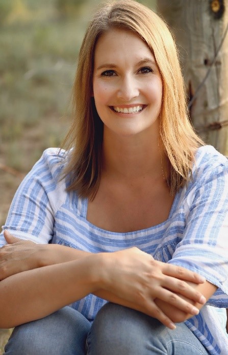 Portrait of a smiling woman with blonde hair, wearing a blue and white striped shirt, sitting outdoors against a tree.