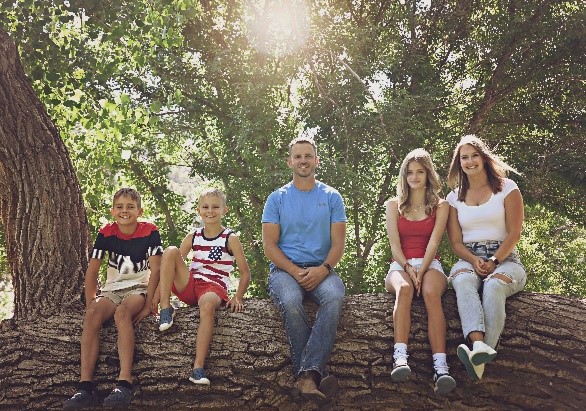A family of five sitting on a tree branch outdoors, with sun rays filtering through the trees.