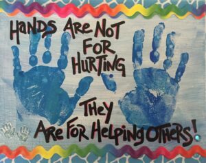 A joyful artwork featuring two large blue handprints and two smaller white handprints on a canvas with a wavy, multicolored border. The message 'Hands Are Not For Hurting, They Are For Helping Others!' is written in bold letters, emphasizing the theme of compassion and assistance. The image signifies the positive impact we can have when we use our hands to support and care for one another.